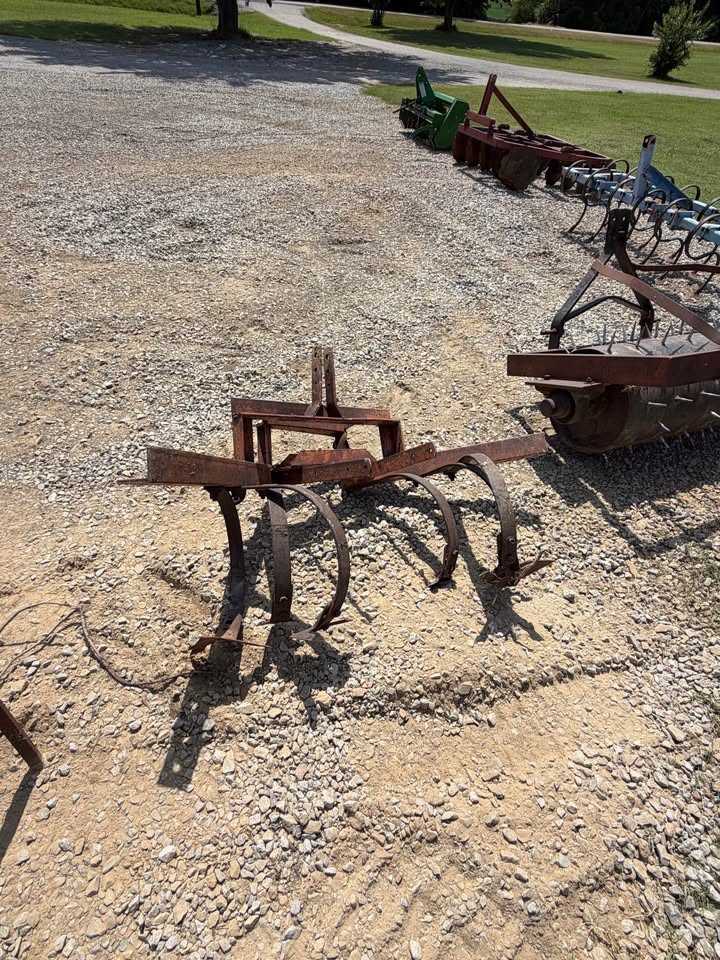 A rusty metal soil tiller or cultivator lying on a gravel lot outdoors, with other farm implements and grassy land in the background.