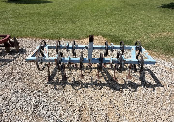 A metal garden or lawn roller with curved black wheels and a light blue frame, resting on a gravel surface in an outdoor yard with grass and trees in the background.