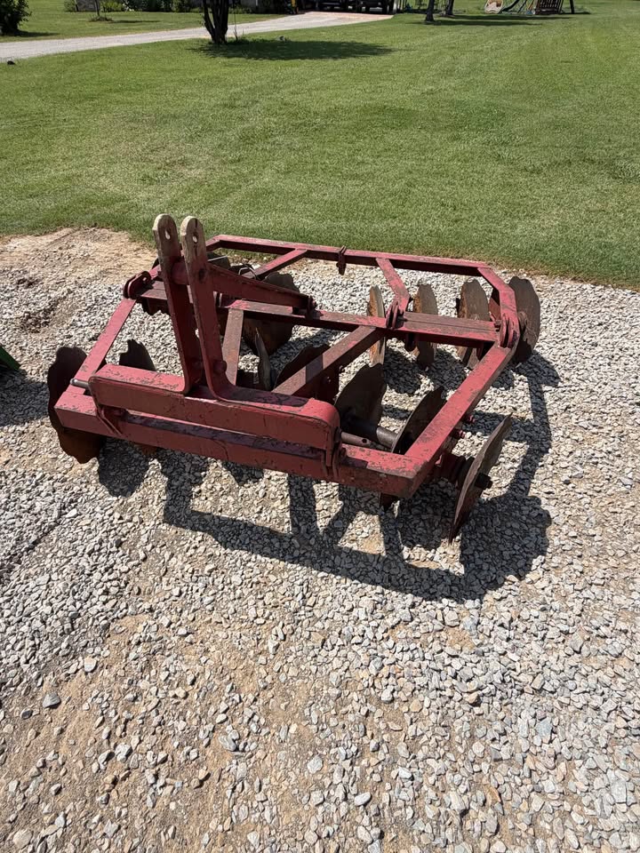 A red metal agricultural soil opener or tillage tool with multiple curved blades, photographed outdoors on a gravel surface with grass in the background.